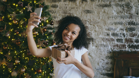 Funny mixed race girl taking selfie pictures on smartphone camera at home near Christmas treeの写真素材