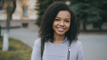Portrait of mixed race curly student girl smiling into camera and laughing at city streetの写真素材