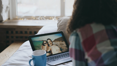 Curly young woman having online video chat with friends using laptop camera while lying on bedの写真素材
