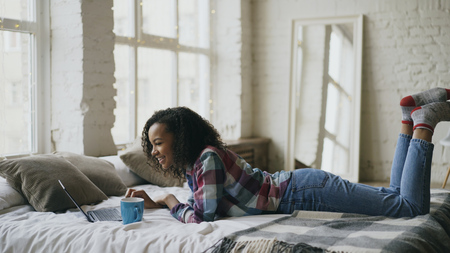 Curly mixed race student girl lauging using laptop for sharing social media lying in bed at homeの写真素材