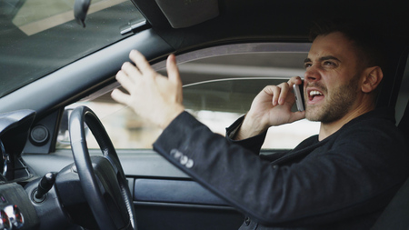 Stressed businessman swearing and talking phone while sitting inside car outdoorsの写真素材