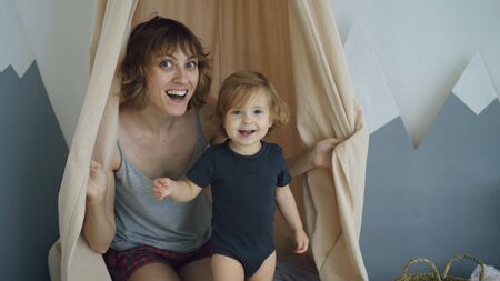 Happy mother with young cute daughter playing and and hide behind curtain at homeの写真素材