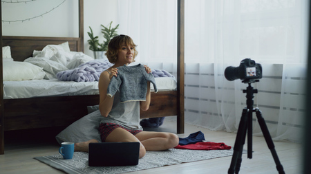 Attractive cheerful woman sitting near bed recording video blog about childrens clothes with dslr camera at homeの写真素材