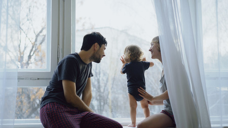 Happy family with young cute daughter sitting on windowsill playing and looking in window at homeの写真素材