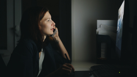 Concentrated woman designer talking phone working in office at night using computer and graphics tablet to finish jobの写真素材