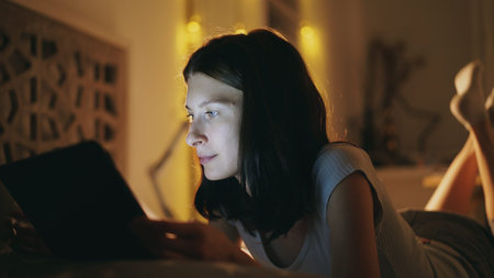 Young smiling woman using tablet computer for sharing social media lying in bed at home at night