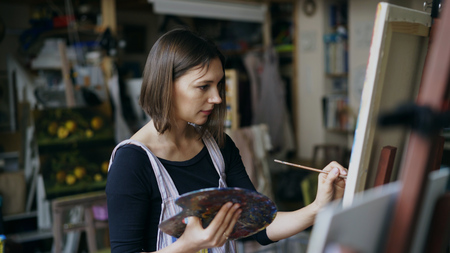 Young woman artist in apron painting picture on canvas in art studioの写真素材