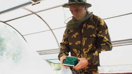 Young beekeeper man smoking bees away from beehive in apiary