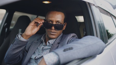 Portrait of serious businessman sitting inside car put of sunglasses and smiling into camera outdoorsの写真素材