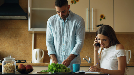 Attractive couple in the kitchen early morning. Beautiful girl chatting phone while her boyfriend cooking breakfastの写真素材