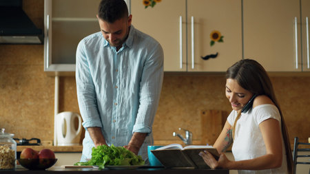 Attractive couple in the kitchen early morning. Beautiful girl chatting phone while her boyfriend cooking breakfastの写真素材