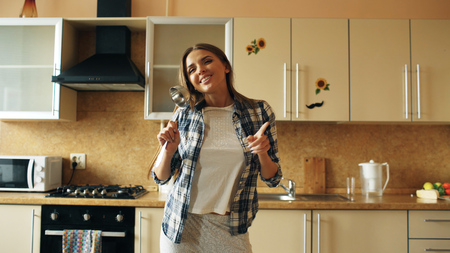 Cheerful young funny woman dancing and singing with ladle while having leisure time in the kitchen at homeの写真素材
