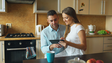 Happy young couple using digital tablet computer while sitting in the kitchen and having breakfast in the morningの写真素材