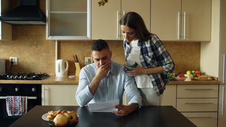 Upset young man reading unpaid bills and hugged by his wife supporting him in the kitchen at homeの写真素材