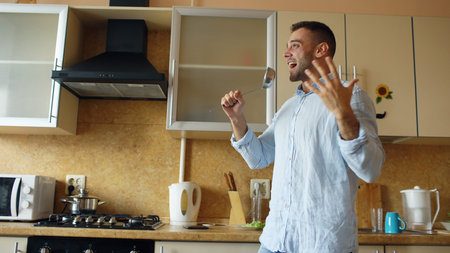 Attractive young funny man dancing and singing with ladle while cooking in the kitchen at homeの写真素材