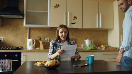Attractive happy couple recieve good news unfolding letter in the kitchen while have breakfast at homeの写真素材
