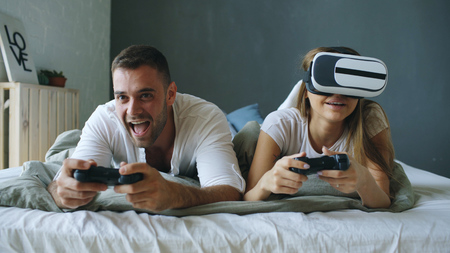 Young couple lying in bed play video games with controller and VR headsetの写真素材