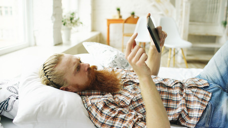 Young bearded man using tablet computer lying on bed in bedroom at homeの写真素材