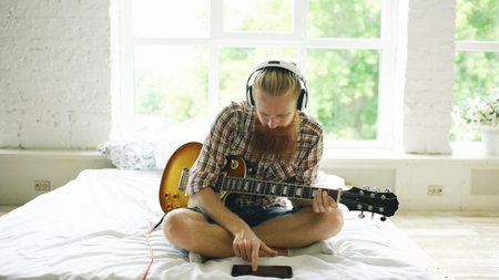 Attractive bearded man in headphones sitting on bed learning to play guitar using tablet computer in modern bedroomの写真素材