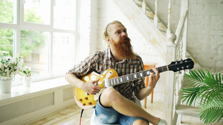 Attractive bearded man sitting on chair learning to play guitar in modern bedroom at homeの写真素材
