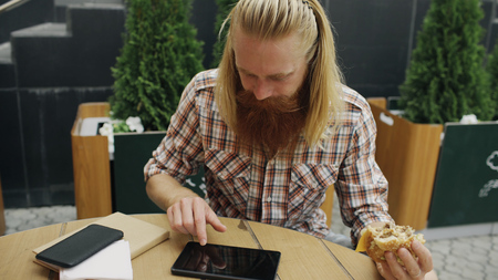 Bearded student man eating burger while surfing tablet computer in street cafe outdoorsの写真素材