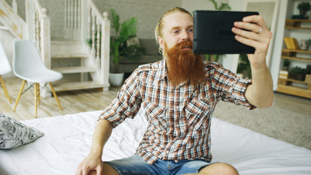 Young bearded man using tablet computer having video chat sitting in bed at homeの写真素材