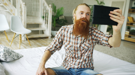 Young bearded man using tablet computer having video chat sitting in bed at homeの写真素材