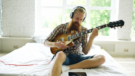 Attractive bearded man sitting on bed learning to play guitar using tablet computer in modern bedroom at homeの写真素材