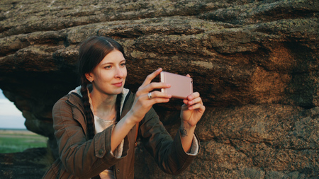 Young tourist woman backpacker photographing landscape on her smartphone camera after hiking on rock at sunsetの写真素材