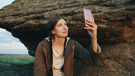 Young tourist woman backpacker photographing landscape on her smartphone camera after hiking on rock at sunsetの写真素材
