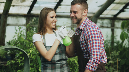 Happy young florist couple in apron have fun while working in greenhouse. Laughing woman spray water in husband faceの写真素材