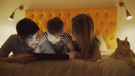 Happy family with little son and funny cat lying in bed at home and using tablet computer for watching cartoon movie before sleepingの写真素材
