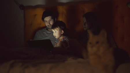 Happy family with little son learning to play tablet computer lying in bed with cat at homeの写真素材