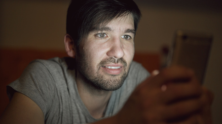 Closeup of Young smiling man using smartphone for surfing social media lying in bed at home at nightの写真素材
