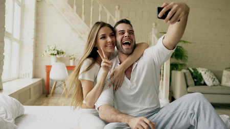 Young beautiful and loving couple take selfie picture on smartphone camera while sitting in bed at the morningの写真素材