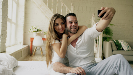 Young beautiful and loving couple take selfie picture on smartphone camera while sitting in bed at the morningの写真素材