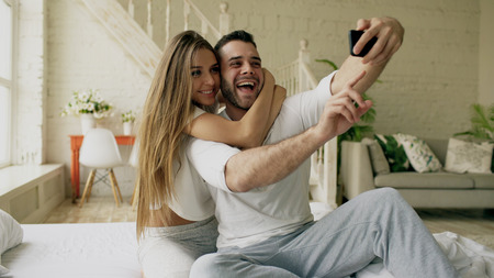 Young beautiful and loving couple take selfie picture on smartphone camera while sitting in bed at the morningの写真素材