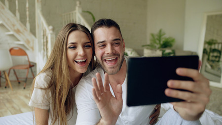 Young cute and loving couple having video chat holding tablet computer and chatting to parents sitting in bed at homeの写真素材