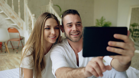 Closeup of Young cute and loving couple having video chat holding smartphone and chatting to friends sitting in bed at homeの写真素材