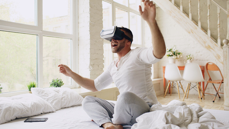 Young cheerful man wearing virtual reality headset having 360 VR video experience while sitting in bed at homeの写真素材
