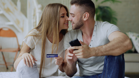 Attractive young couple with smartphone and credit card shopping on the internet sit on bed at homeの写真素材