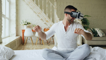 Young cheerful man wearing virtual reality headset having 360 VR video experience while sitting in bed at homeの写真素材