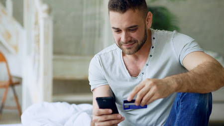 Attractive young man with smartphone and credit card shopping on the internet sit on bed at homeの写真素材