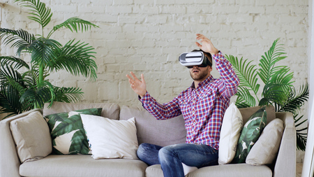 Young cheerful man wearing virtual reality headset having 360 VR video experience while sitting on couch in living room at homeの写真素材