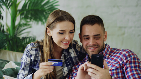 Attractive young couple with smartphone and credit card shopping on the internet sit on couch in living room at homeの写真素材