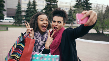 Two cute african american woman taking selfie with shopping bags and smiling. Friends have fun after...