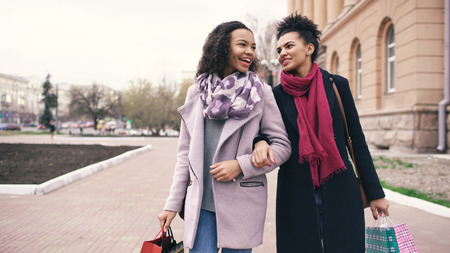 Two attractive mixed race women with shopping bags talking and walking down the street. Girlfriends have fun after visiting mall saleの写真素材