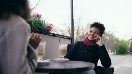 Two mixed race women sitting at the table in the street cafe using smartphone and talkingの写真素材