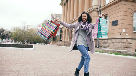Attractive mixed race girl dancing and have fun while walking down the street with bags. Happy young woman walking after shopping on mall saleの写真素材