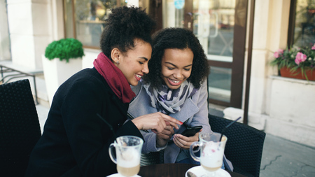 Two attractive mixed race female friends sharing together using smartphone in street cafe outdoorsの写真素材